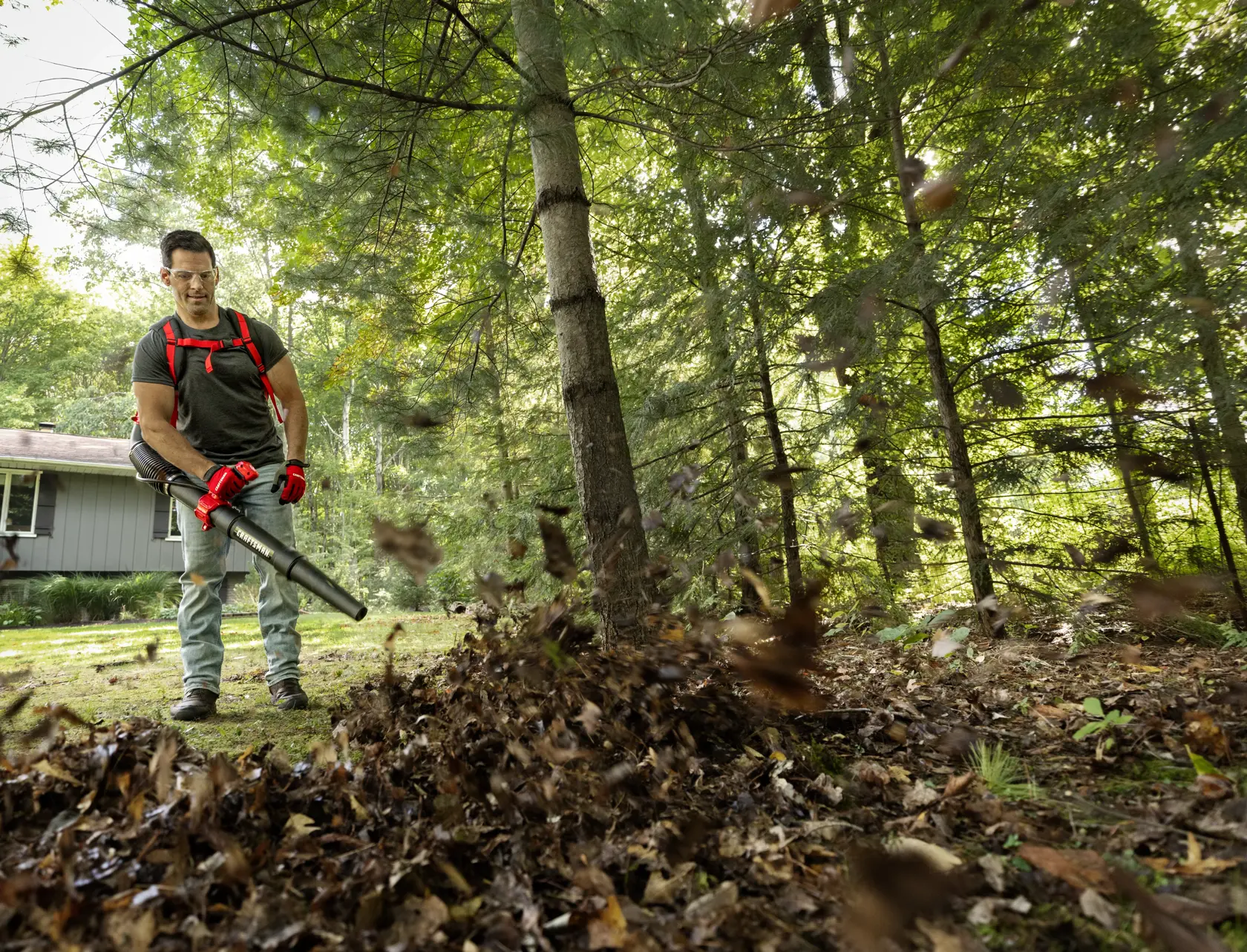 Photo of person using CRAFTSMAN backpack leaf blower CM41AR51BP593 in yard, blowing leaves with house and trees in background.