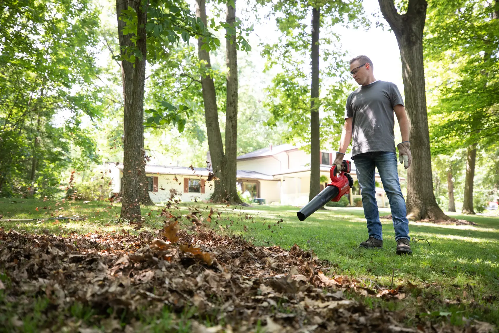 Photo of person using CRAFTSMAN cordless leaf blower, SKU CMCBL730P1, clearing leaves in a grassy yard with trees.