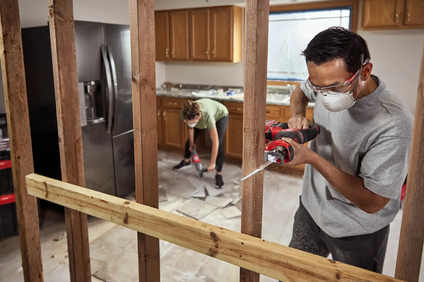 Two people using power tools to renovate a kitchen; one person is cutting through a wooden stud with a reciprocating saw, and another is working on removing floor tiles. The scene shows an unfinished area with wooden framing and construction debris.