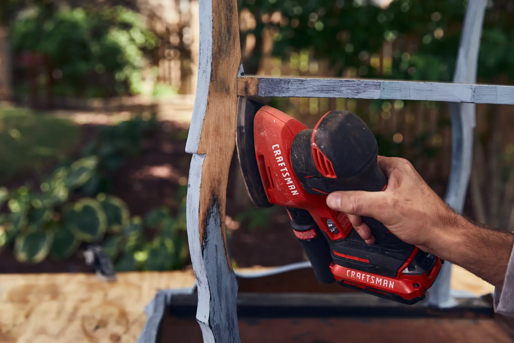Photo of a hand using CRAFTSMAN CMCW221B cordless detail sander on a wooden furniture piece outdoors.