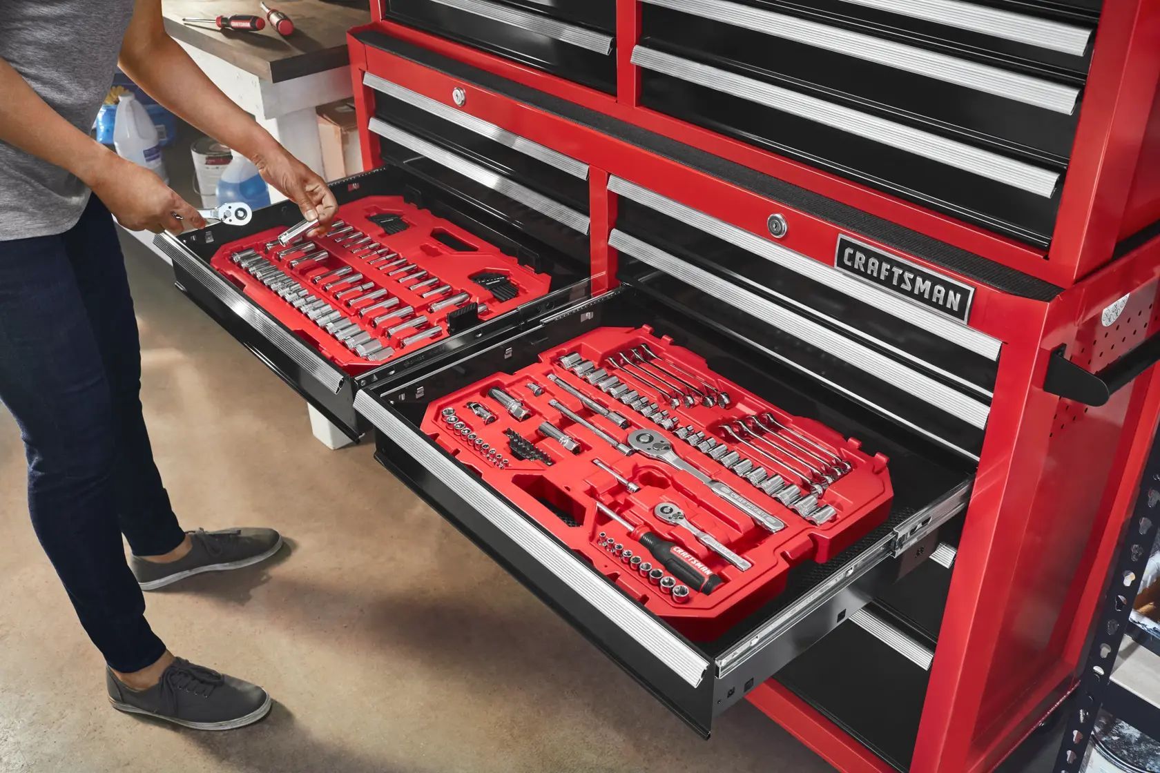 A person is accessing red tool trays filled with assorted hand tools and socket sets stored in the drawers of a red Craftsman tool chest.
