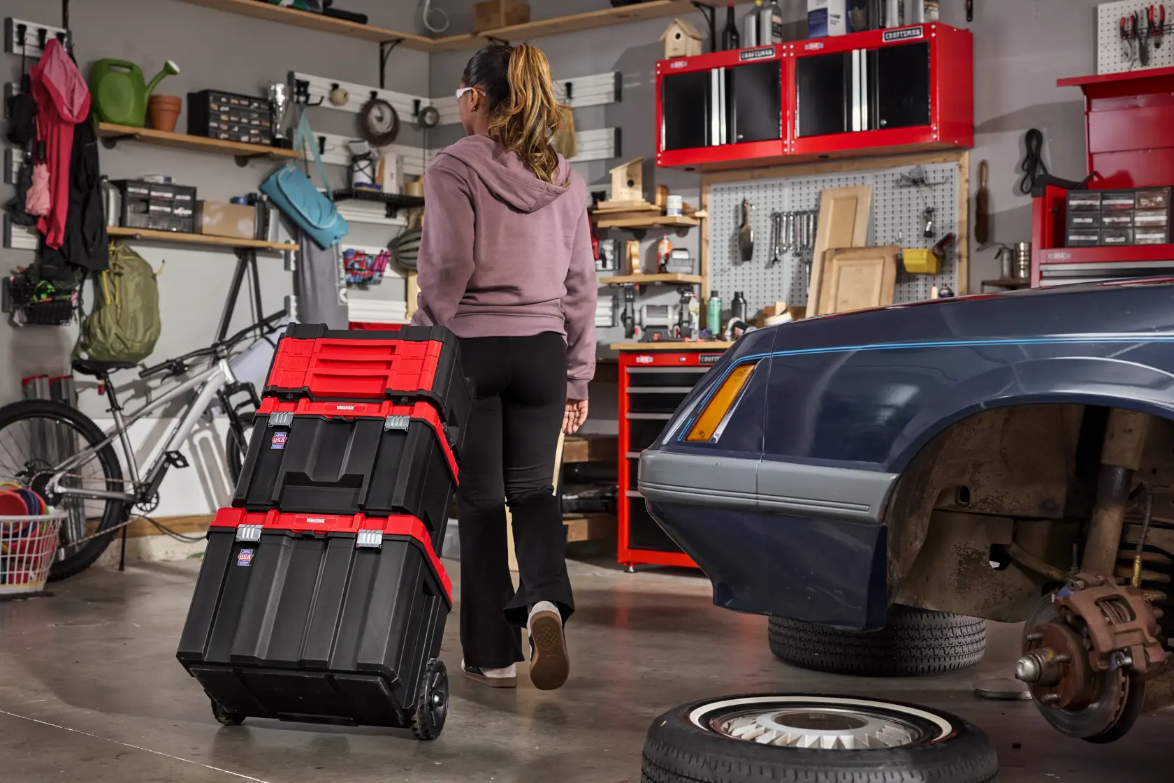 A person is pulling a stack of black and red rolling toolboxes in a garage. The garage contains shelves with various tools and supplies, a bicycle, and a partially repaired car with a detached tire.