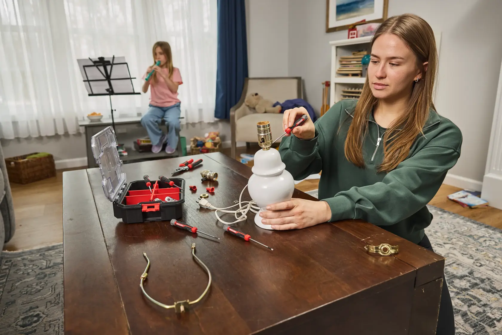 A person is repairing a white table lamp on a wooden table using hand tools from a CRAFTSMAN toolbox. Several screwdrivers and lamp parts are scattered on the table. In the background, another person is playing a wind instrument and reading sheet music. The setting is a cozy, well-lit living room.