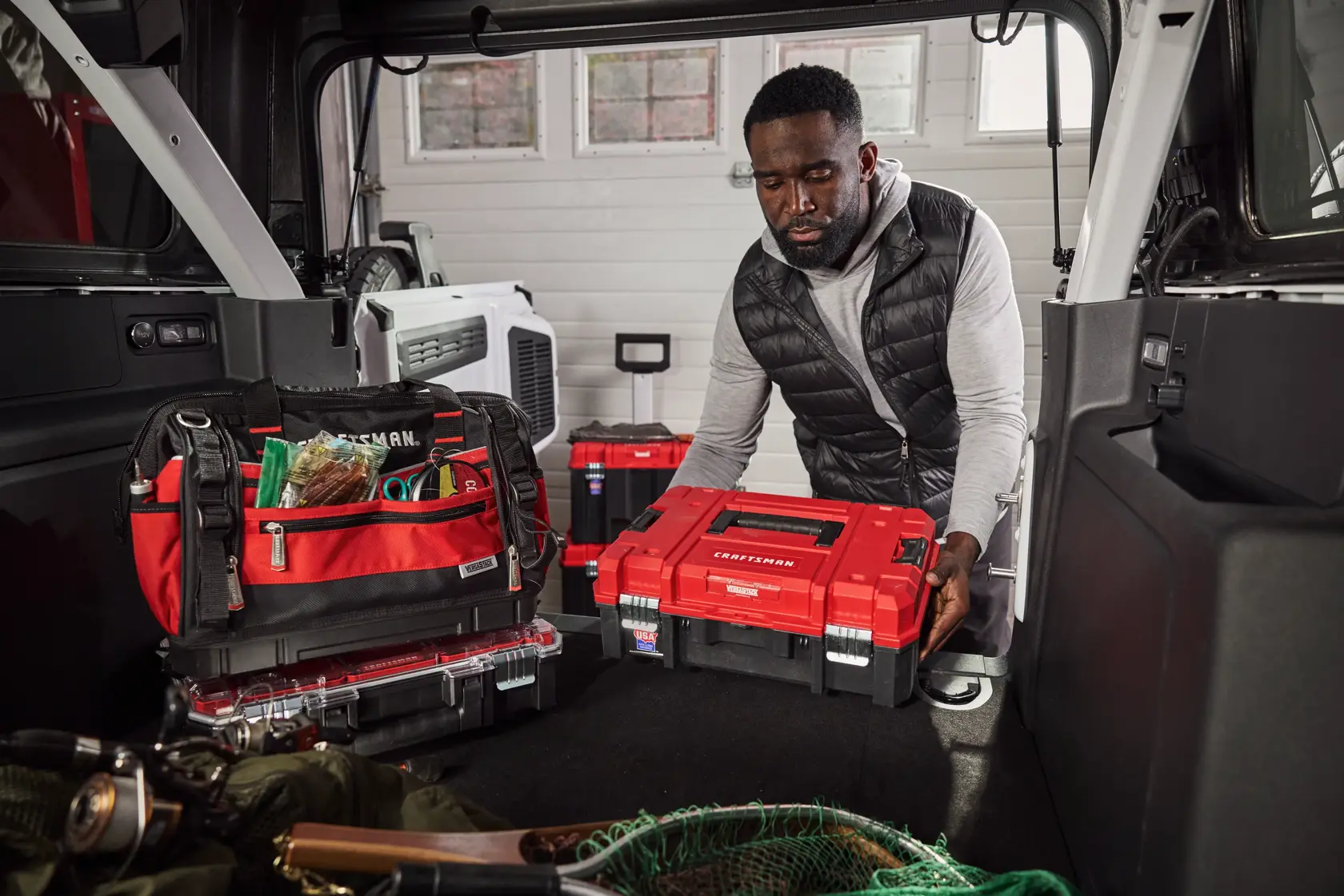 A person places a red Craftsman toolbox in the back of a vehicle, which is filled with various red and black Craftsman tool bags and fishing equipment. The scene appears to be inside a garage or workshop with windows in the background.