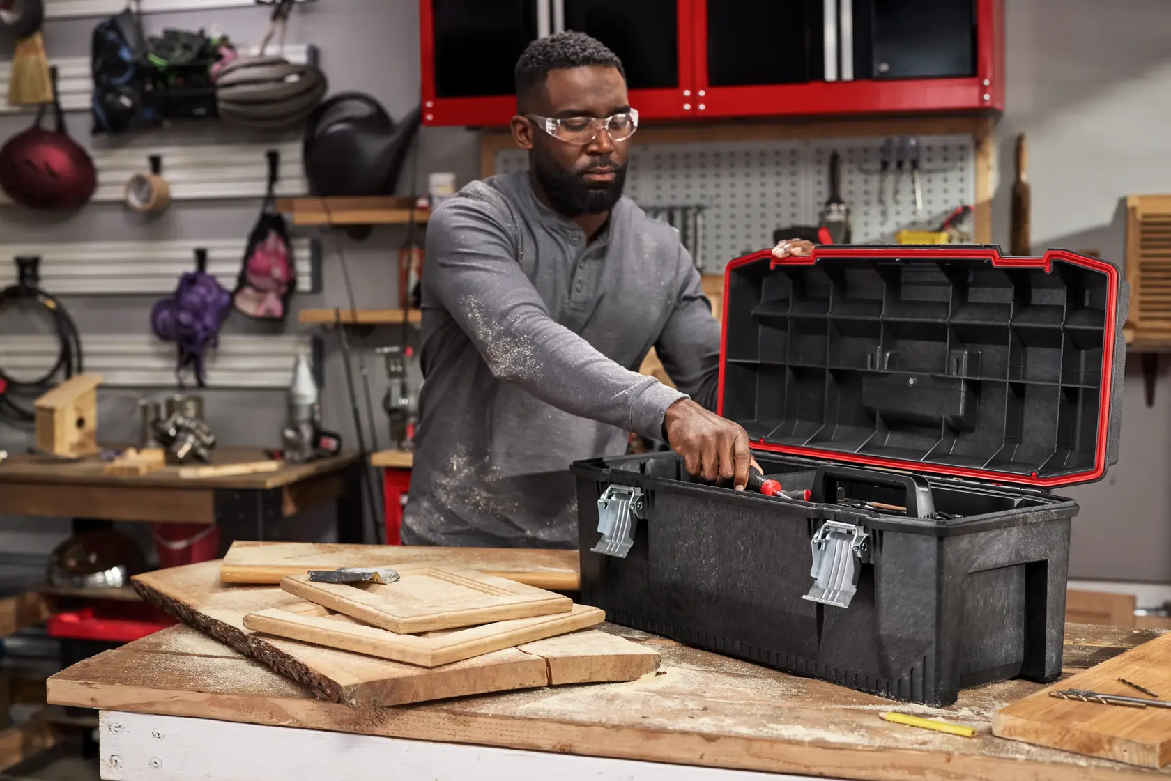 A person standing in a workshop opens a large black toolbox on a workbench. Various tools and wooden boards are visible on the bench, with shelves and equipment in the background.