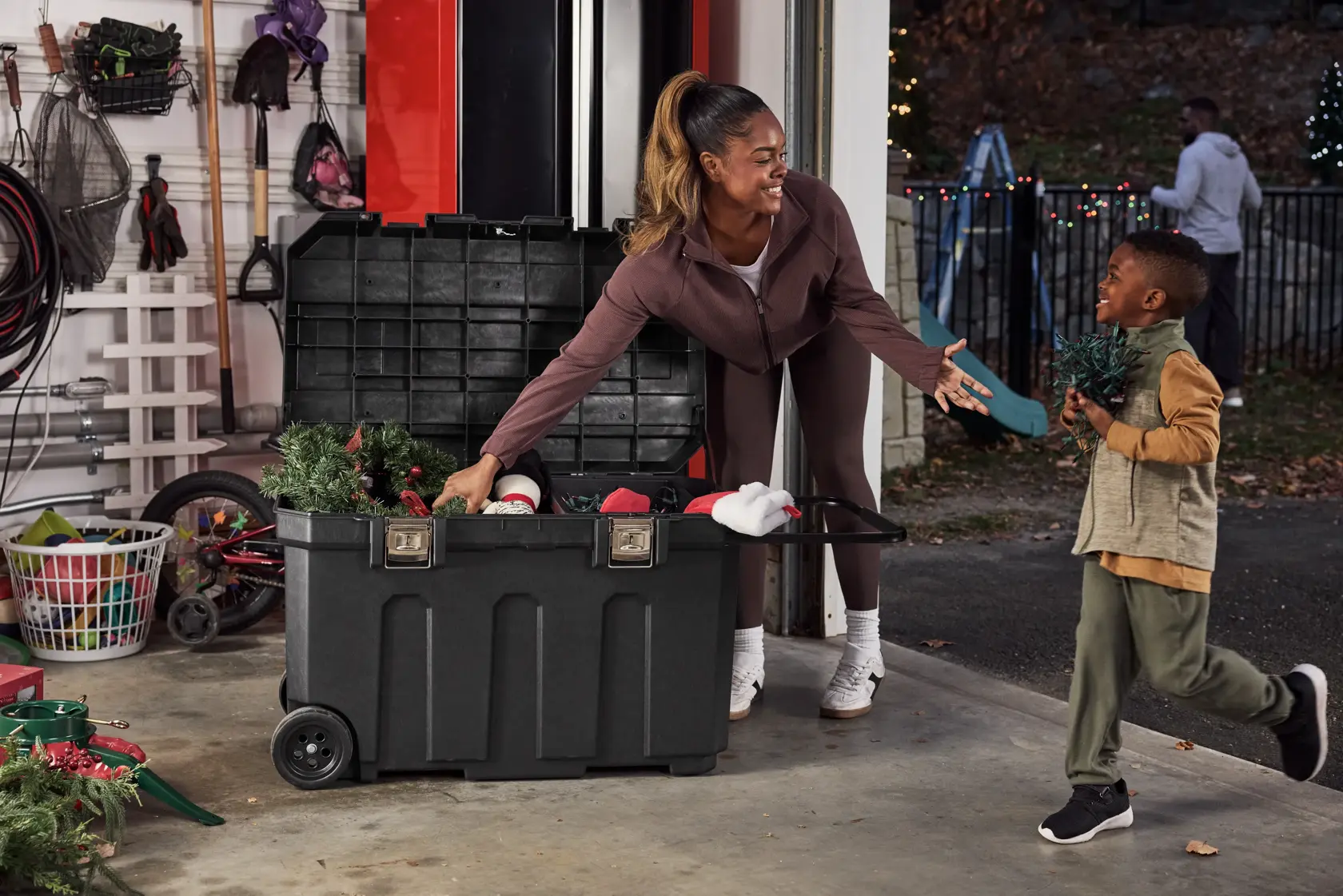 A large black storage trunk with wheels is open in a garage, containing holiday decorations such as wreaths and ornaments. A woman is reaching for decorations in the trunk while a child hands her a small holiday item. Various storage and gardening tools are visible in the background.