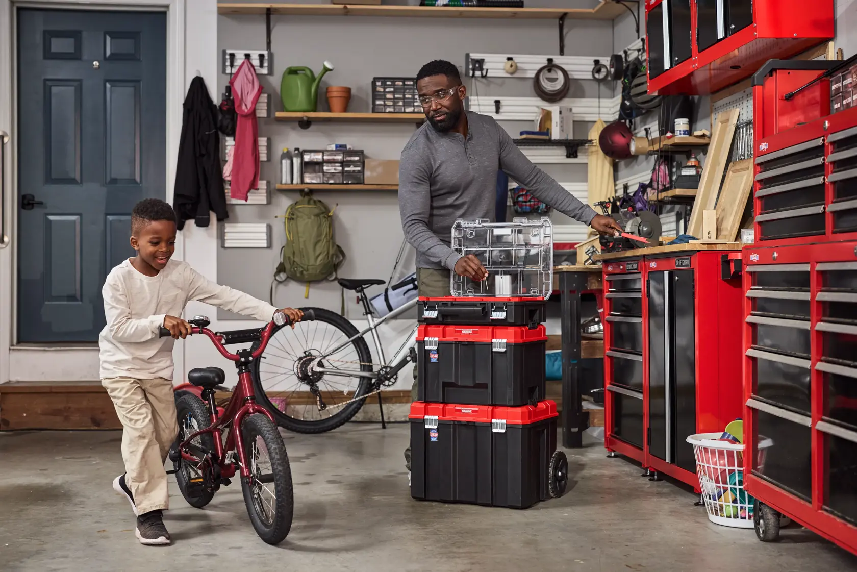 A garage scene showing a person organizing stackable black and red toolboxes with clear compartments. A child walks pushing a red bicycle. The garage contains various tools, cabinets, a laundry basket, and shelves with assorted items in the background.