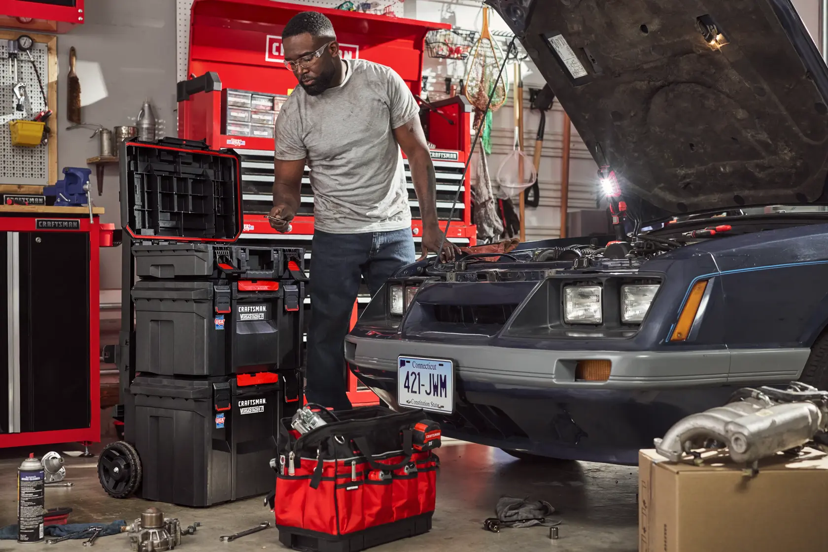 A person working in a garage is using various Craftsman tool storage boxes and a red tool tote, with a car parked nearby that has its hood open for maintenance. Several tools and automotive parts are scattered around the workspace. Shelves and tool cabinets are visible in the background.
